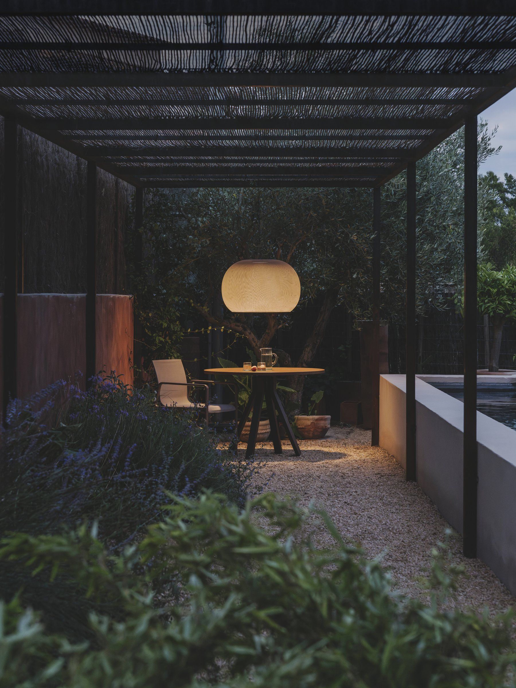 Tuin onder een pergola bij schemer, met een grote ronde hanglamp boven een tafel met stoelen tussen de lavendel