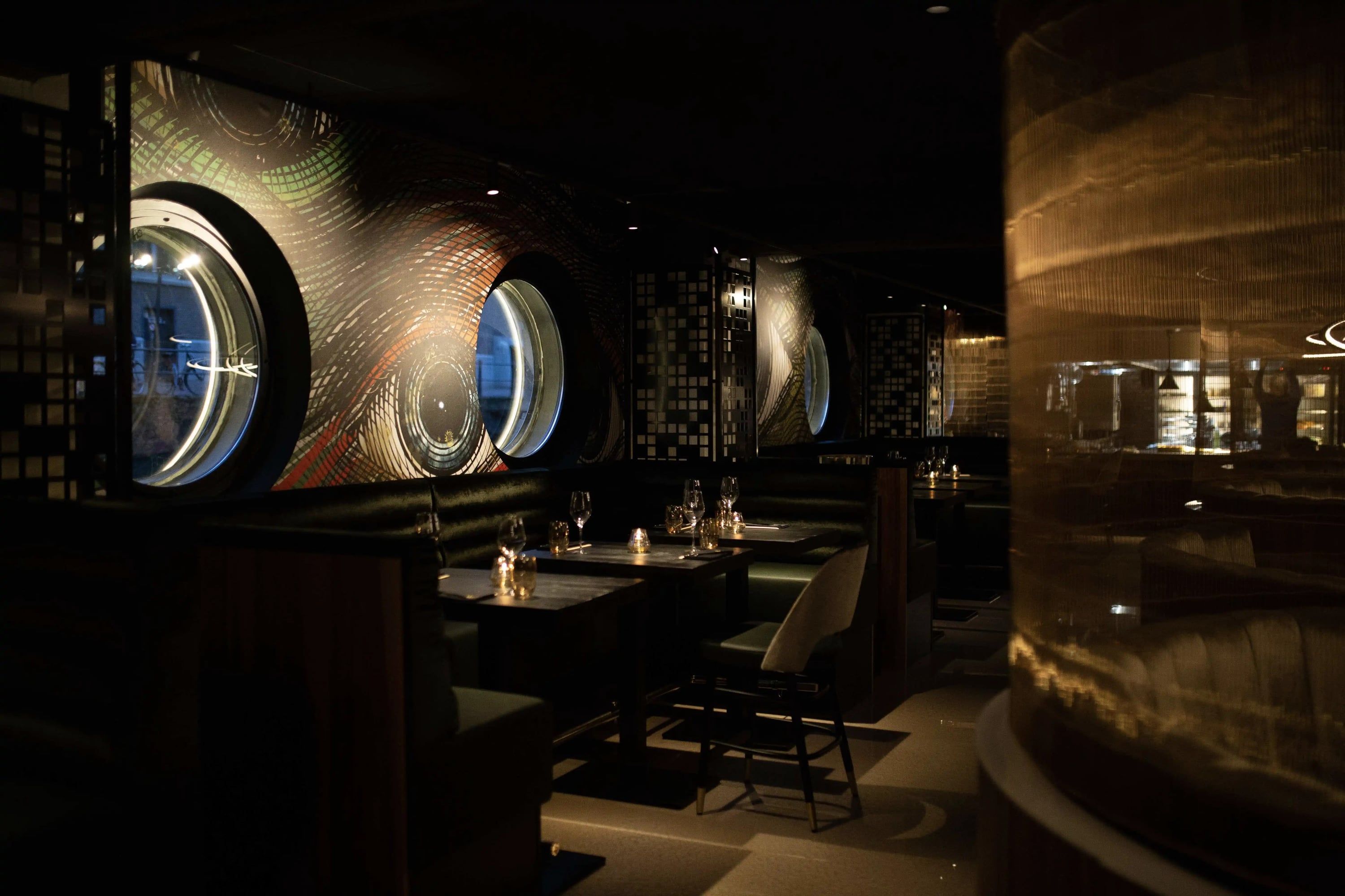 Restaurantzaal met ronde patrijspoortramen en goudkleurige gordijnen in gedempt licht