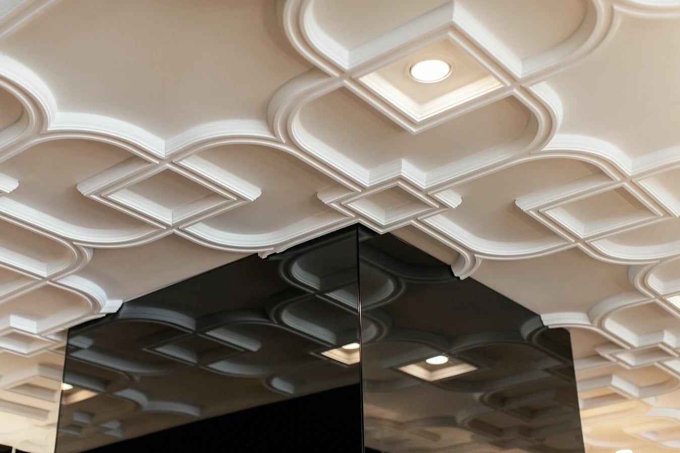 Wit ornamentplafond met vierkante inbouwspots, gespiegeld in een donker vlak