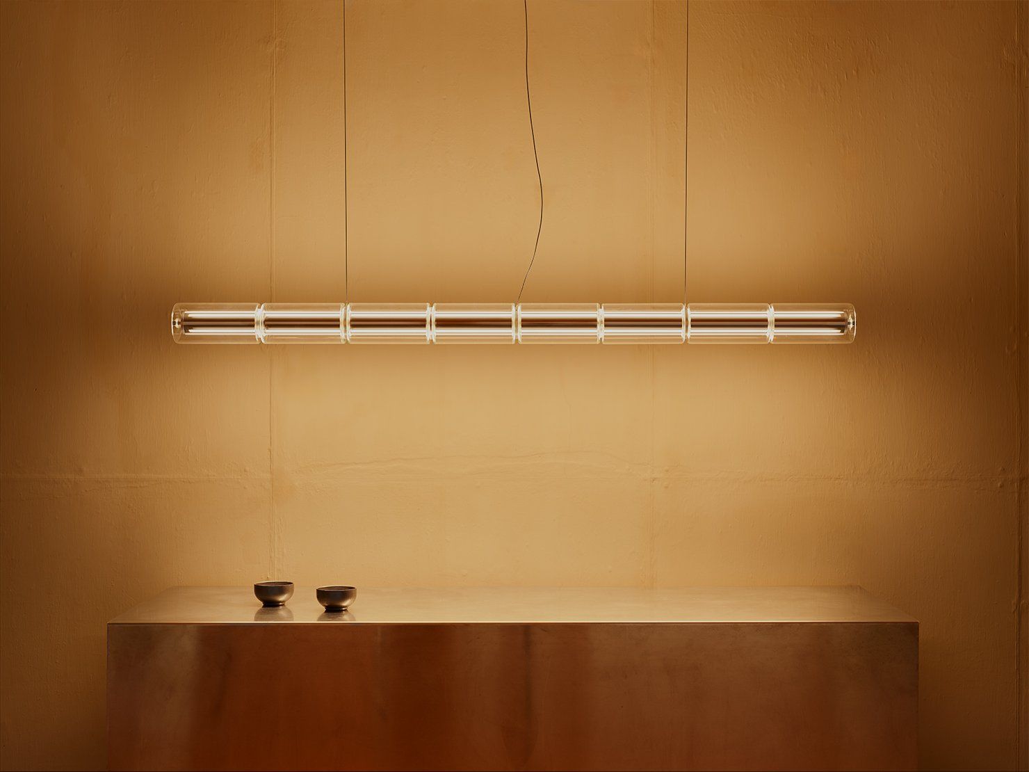 Lange glazen hanglamp met een warme gloed boven een koperkleurig dressoir tegen een okerkleurige wand
