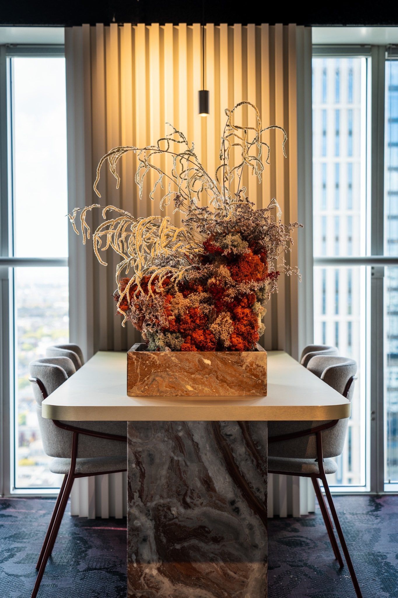 Vergadertafel met marmeren poot en een bloemstuk van droogbloemen voor een verlichte lamellenwand tussen hoge ramen