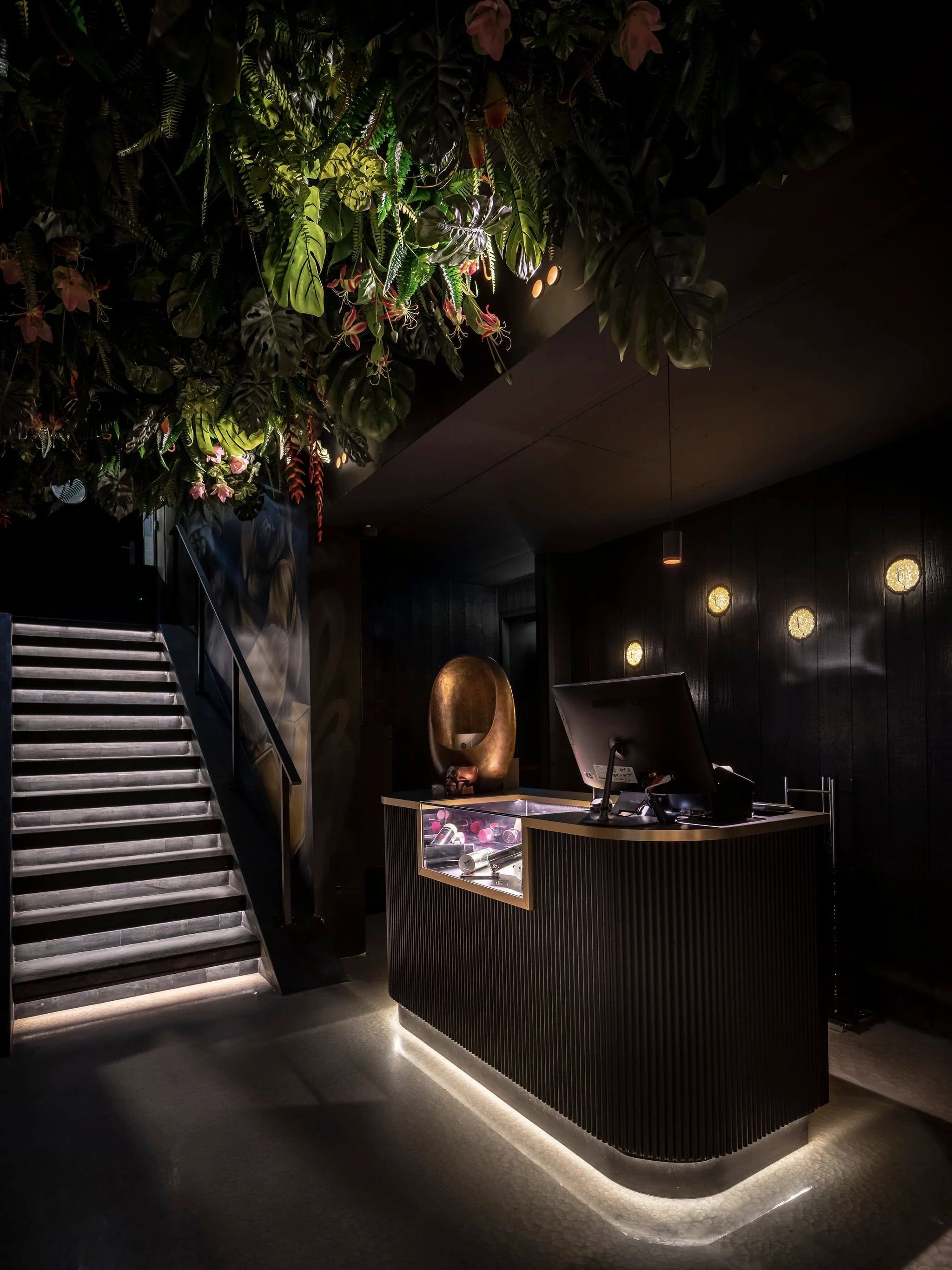Entree met verlichte trap en een balie met een lichtlijn, onder een plafond van planten
