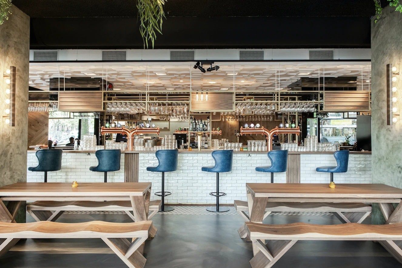 Bar met witte tegels en blauwe barkrukken, met houten picknicktafels ervoor