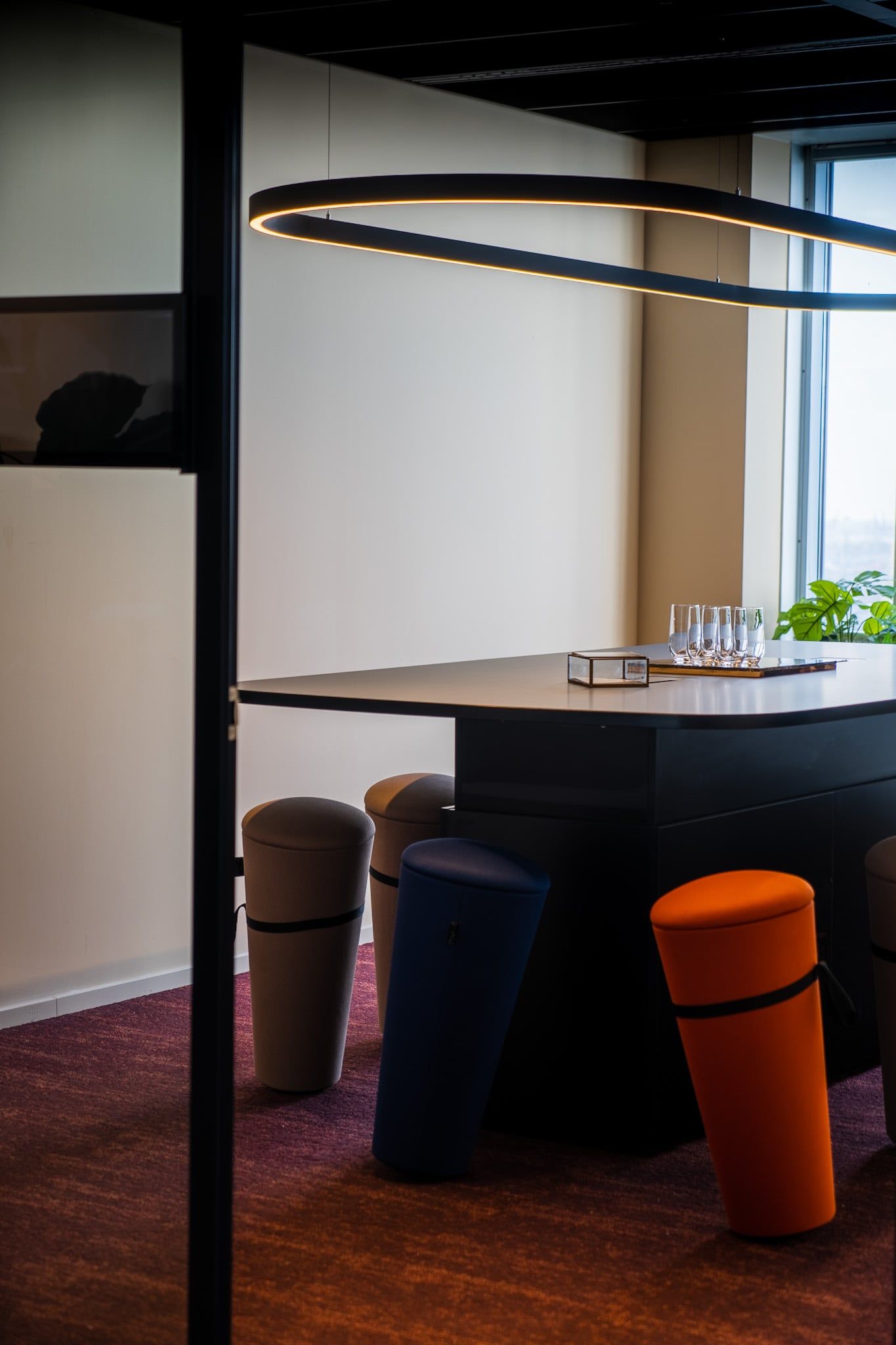 Ovale hanglamp boven een hoge tafel met kleurige zitkrukken in een overlegruimte