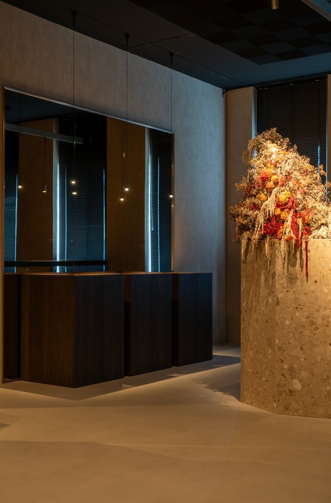 Zuil van terrazzo met een bloemstuk van droogbloemen naast donkerhouten kasten, voor een spiegelwand met kleine hanglampjes